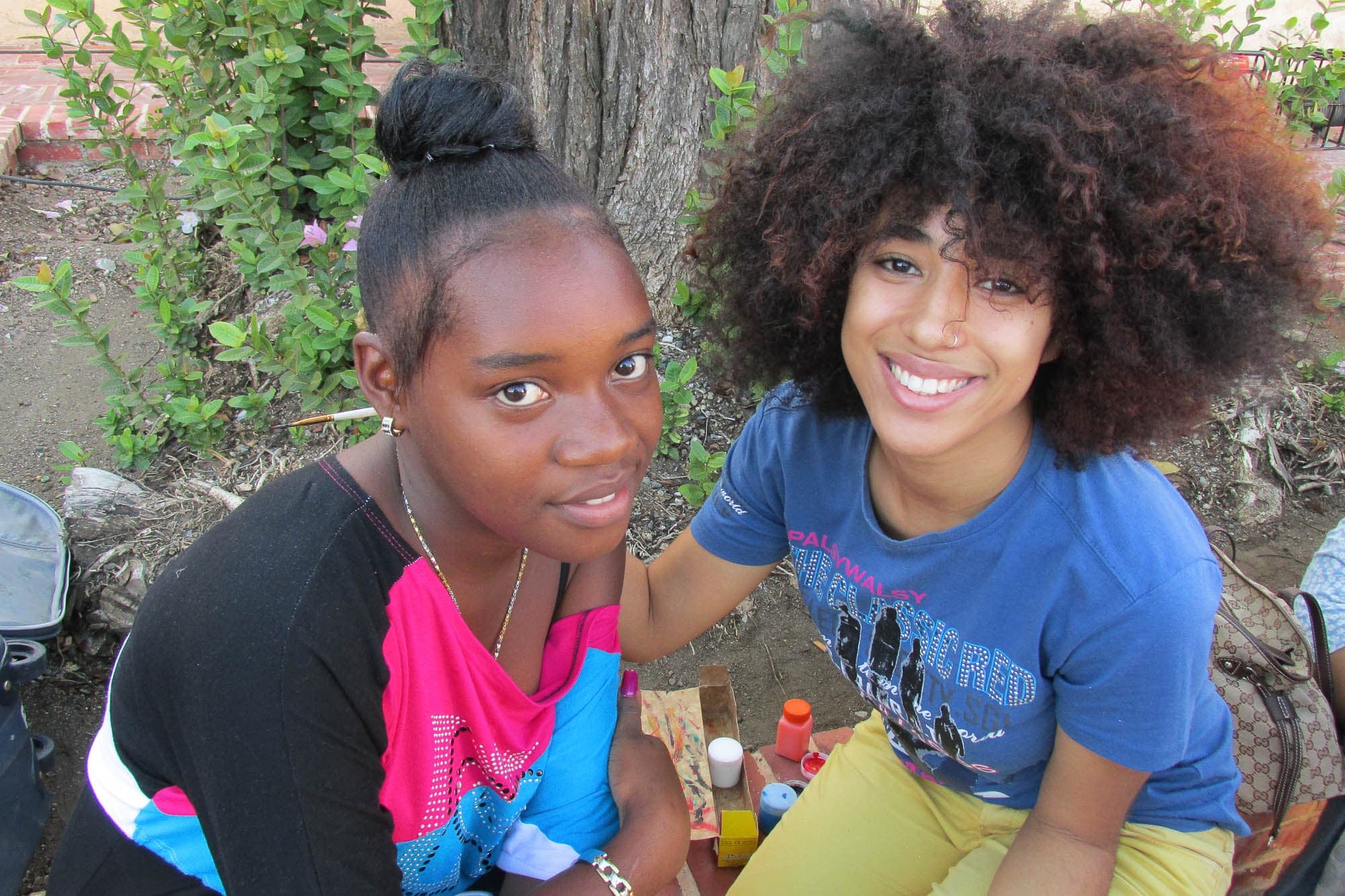 Photograph of two smiling youth sitting outside. One is dark skinned with their black hair in a straight-ironed bun, wearing a black, pink, and blue shirt hanging off one shoulder. The other is medium-dark skinned with dark, curly, chin-length natural hair, wearing a blue shirt and yellow bottoms.