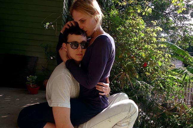 Photograph of a light-skinned person wearing purple clutches the head of a light-skinned person wearing all white and sunglasses while sitting on their lap facing them on the top step of a green house’s porch
