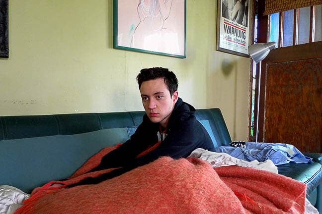 Photograph of a light-skinned person with short dark hair wearing a black hoodie sitting on a couch with a fuzzy orange blanket