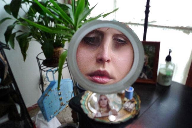 Photograph of a distorted reflection of a light-skinned face with pink lips in a makeup mirror. The mirror is in front of a window with several houseplants in the corner.