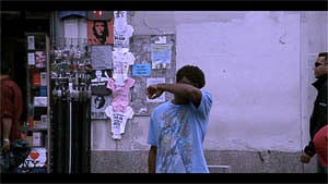 A street scene with a person in a blue shirt covering their face, with posters and advertisements in the background.