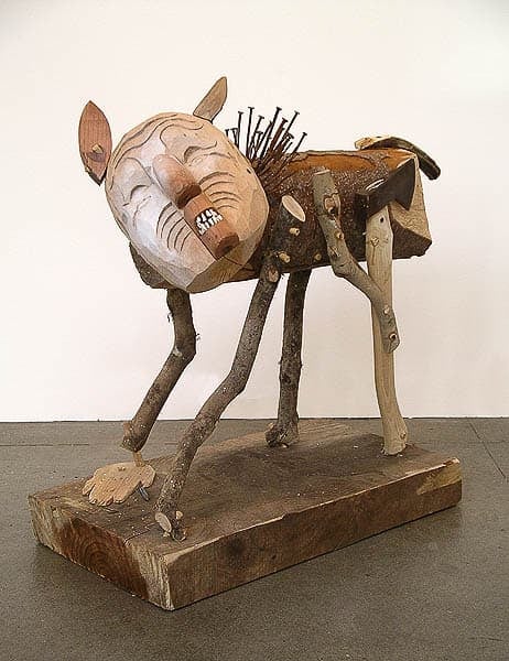 A wooden sculpture resembling a fantastical animal, constructed from branches and pieces of wood. The figure has a carved face with a grimacing expression, nails protruding from its back, and stands on a wooden base.