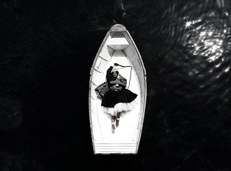 A black and white photograph of a woman lying in a small rowboat on dark water. The woman is dressed in a patterned top and a full skirt, looking relaxed with one arm behind her head and the other holding a paddle.