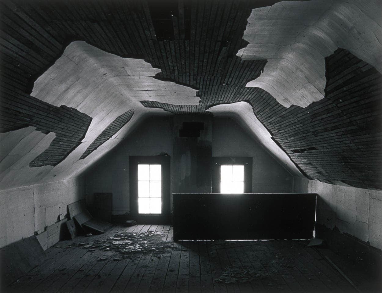 Black and white photograph of an abandoned attic with a peeling wooden ceiling and two windows letting in dim light.
