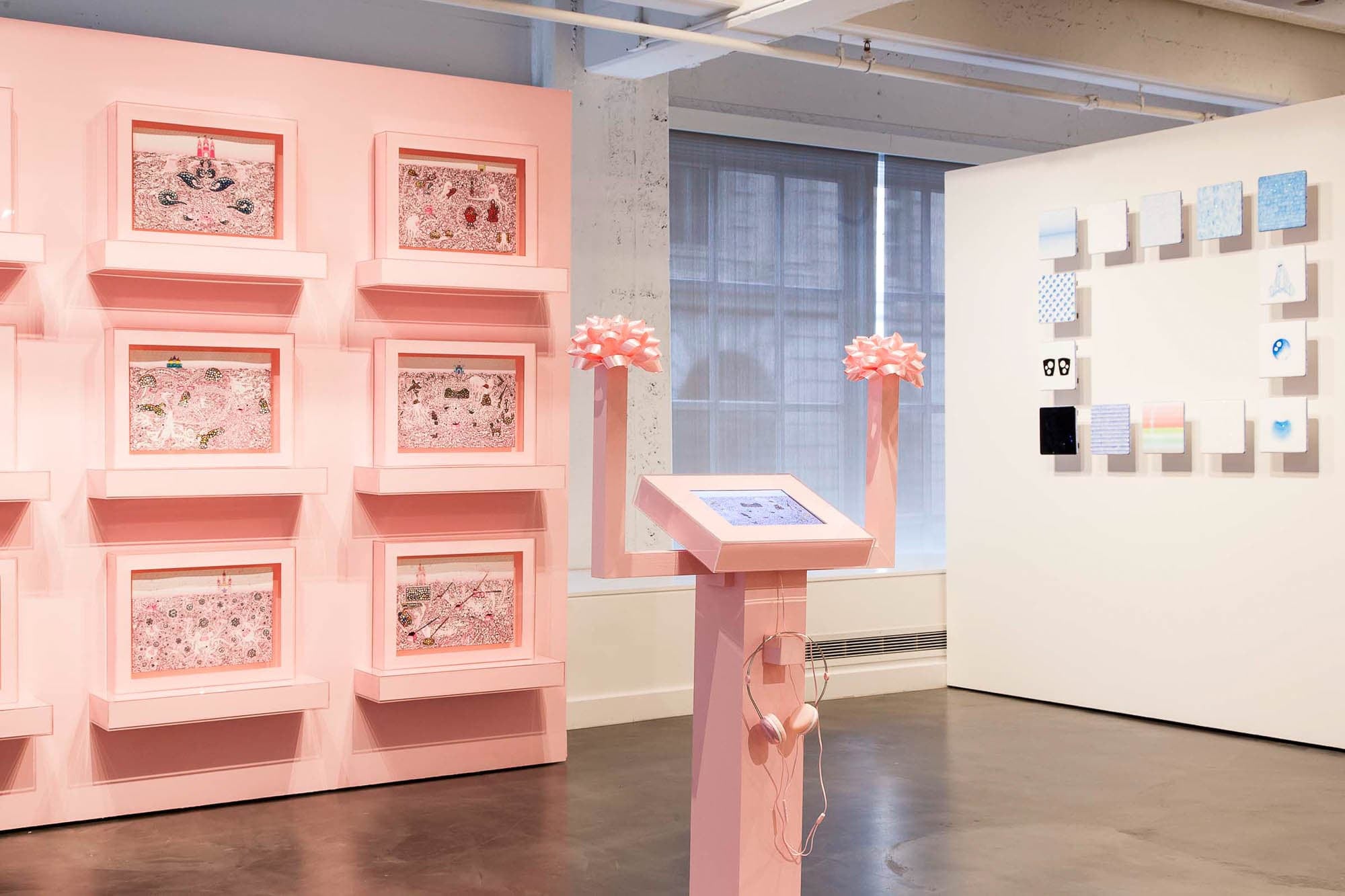 A gallery installation view featuring pink-framed artworks on walls, a central pink pedestal with a touchscreen, and a series of small artworks arranged in a grid on the right wall.