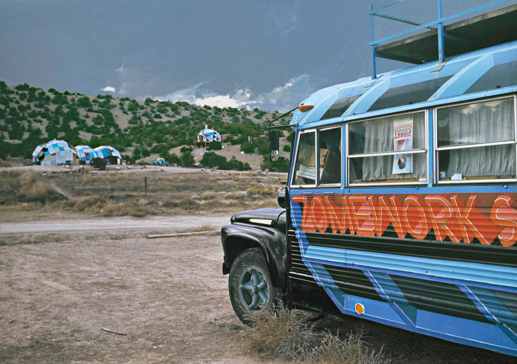 A photograph of a brightly painted bus with the word "Zomeworks" parked in a desert landscape, with geodesic dome structures visible in the background under a cloudy sky.
