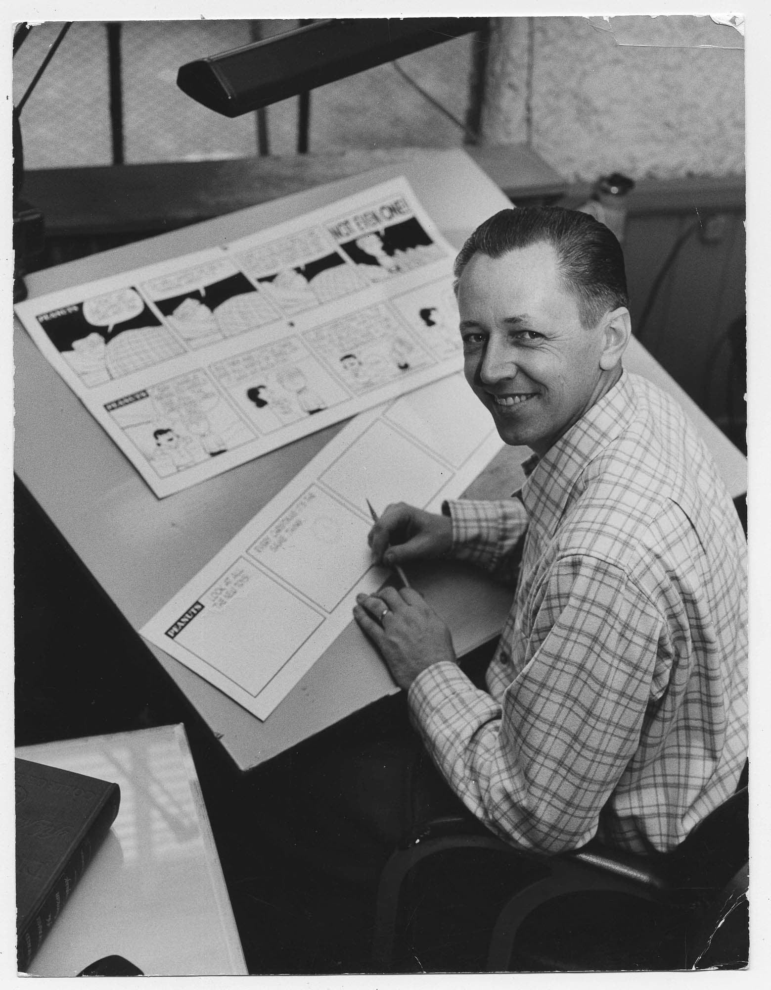 A black and white photo of a smiling man seated at a desk, drawing a comic strip. He is wearing a checkered shirt and looking towards the camera.