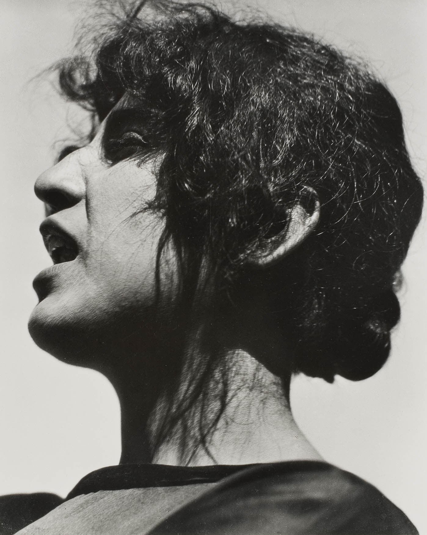 A dramatic black and white photograph of a woman with wavy hair and an expressive face, captured in profile against a light background.