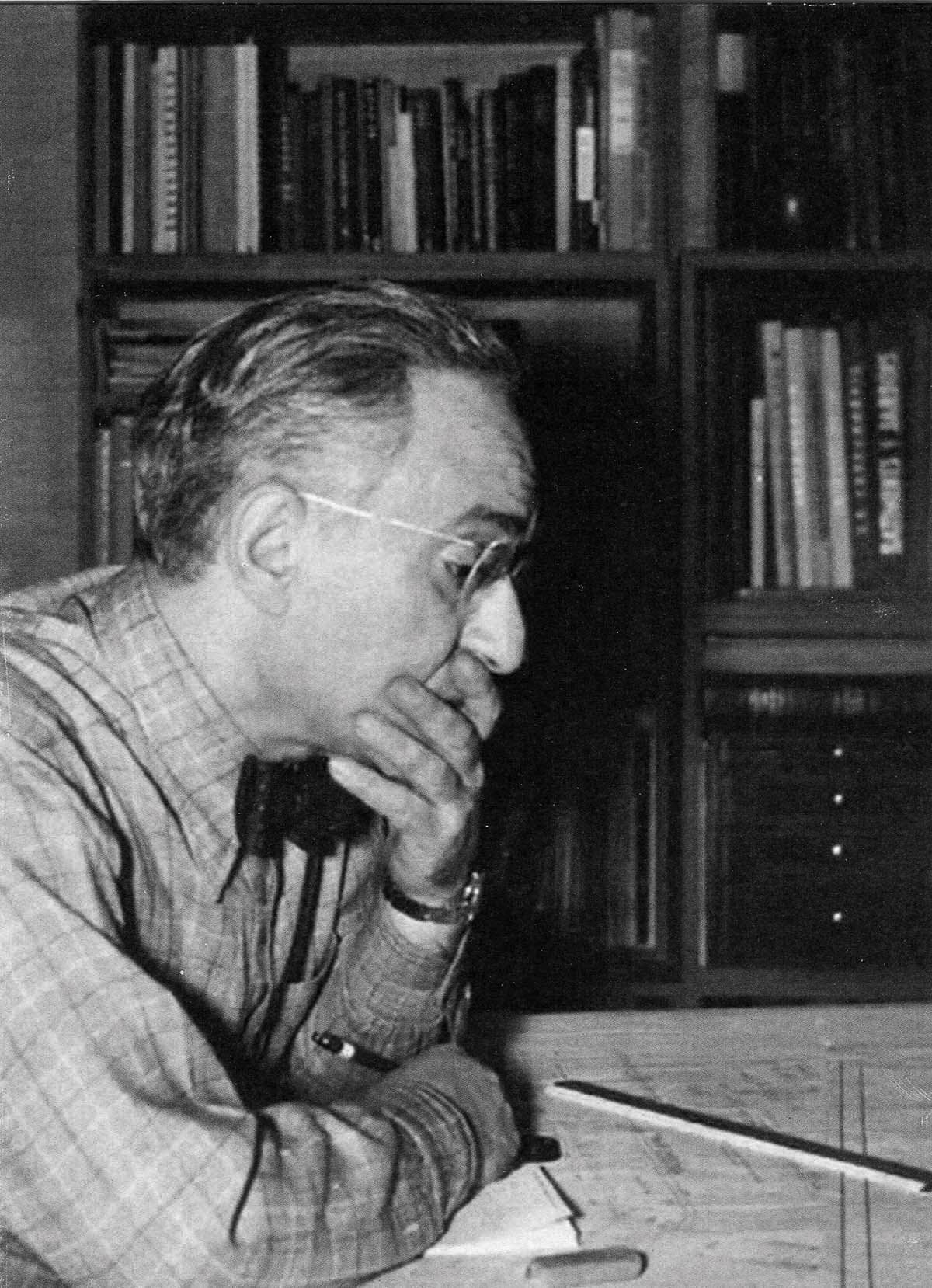 Black and white photograph of a man with glasses, deep in thought, sitting at a desk with bookshelves in the background.