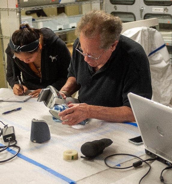 Two individuals in a workshop, one man examining a ceramic piece with a tool, and a woman taking notes beside him.