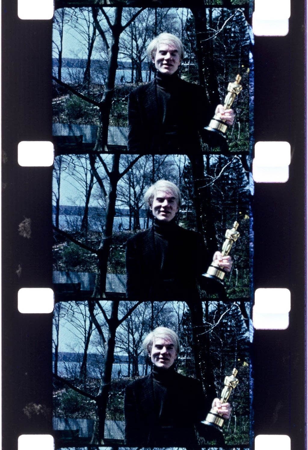 Three grainy color film frames of Andy Warhol, a light-skinned man with unkempt white hair, holding a gold statue. He is wearing black and standing in front of trees and a lake.