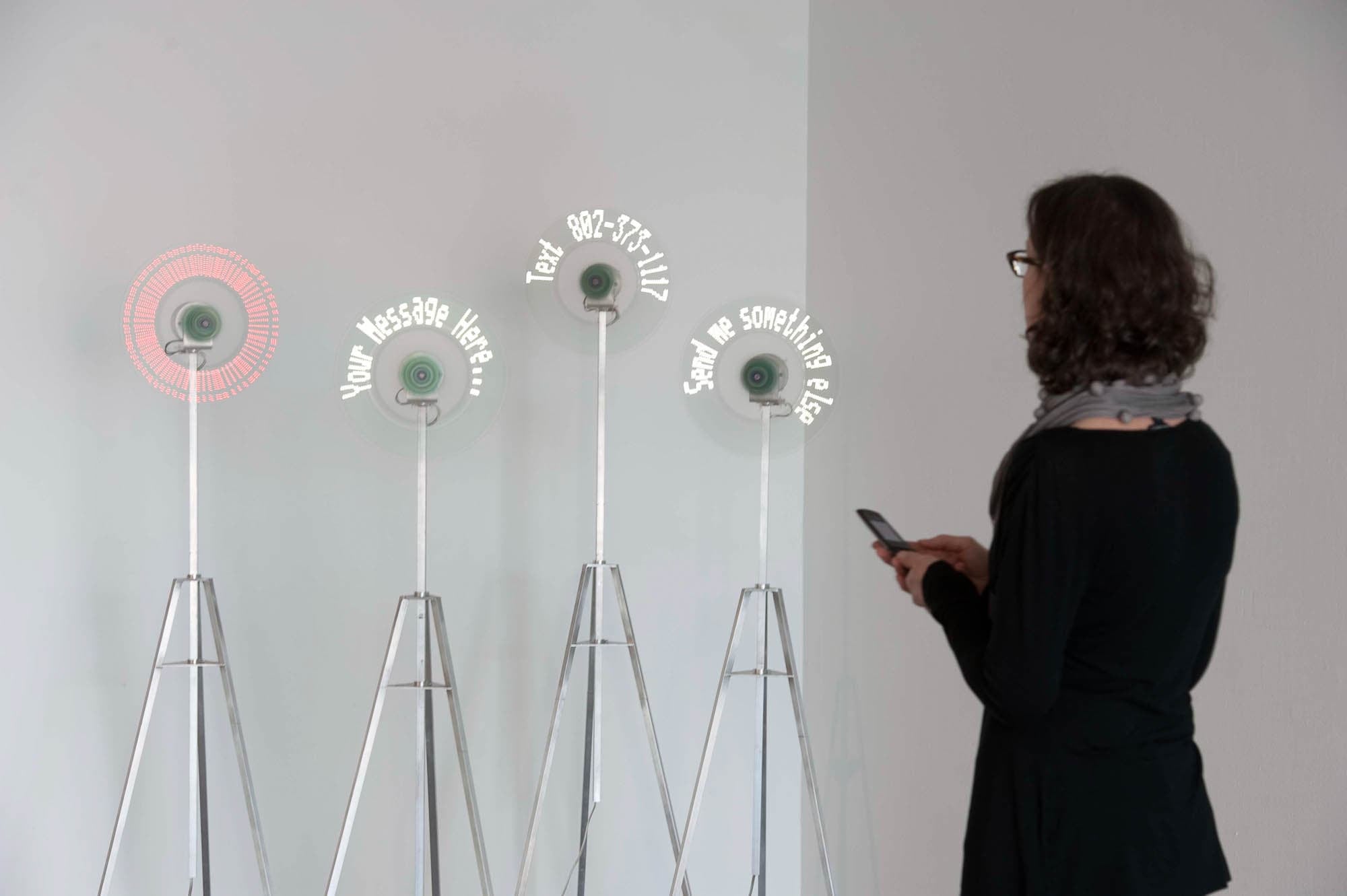 A person with a cell phone standing in front of 4 spinning LED displays. One has created a red circle, the others read, “Your Message Here….” “Text 802-373-1117,” and “Send me something else”