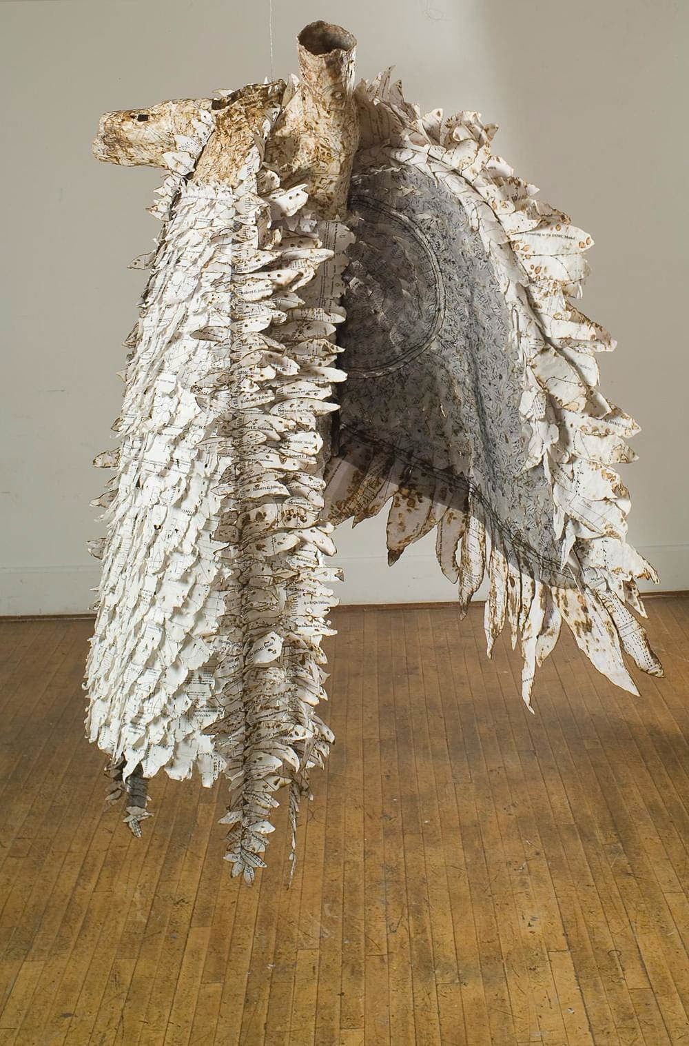 Hanging sculpture of headless, armless body with large white wings wearing a long apron covered with white paper feathers