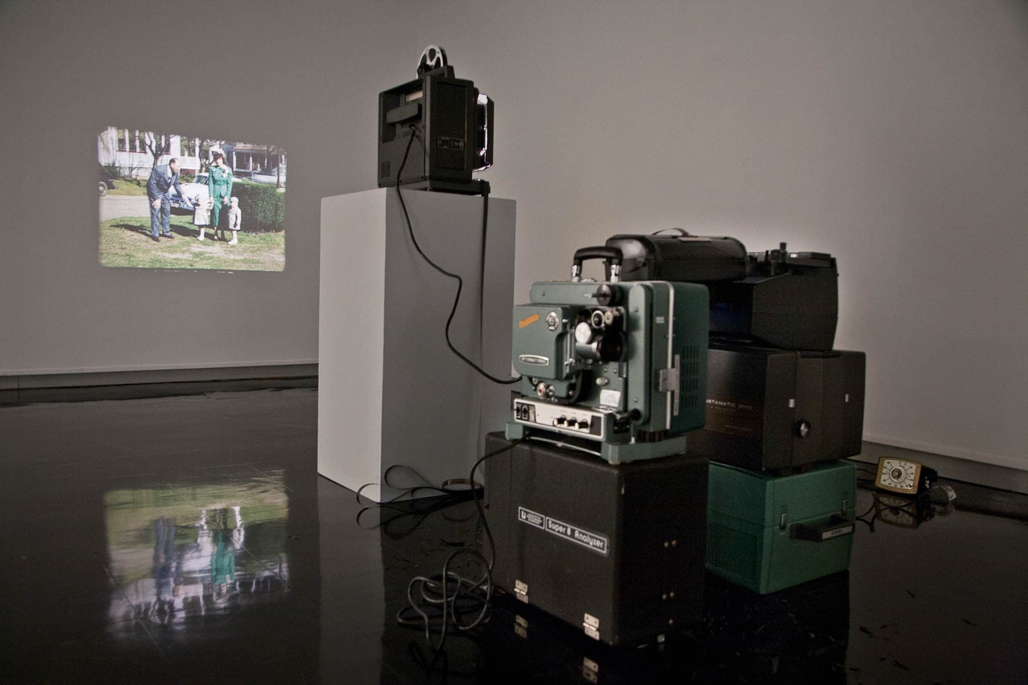 Photograph of installation with a film projector projecting an image of a family on a wall, which also reflects on the floor. A series of green-gray and dark brown cases are stacked behind it.