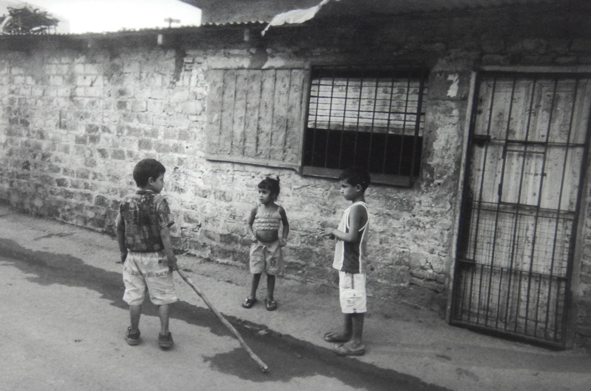 Black and white photo of three children standing on a street outside a cinder block building with a barred window and door