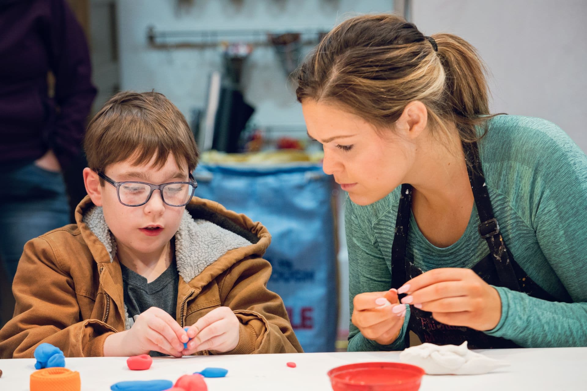 A child in glasses uses modeling clay while a college student assists them.