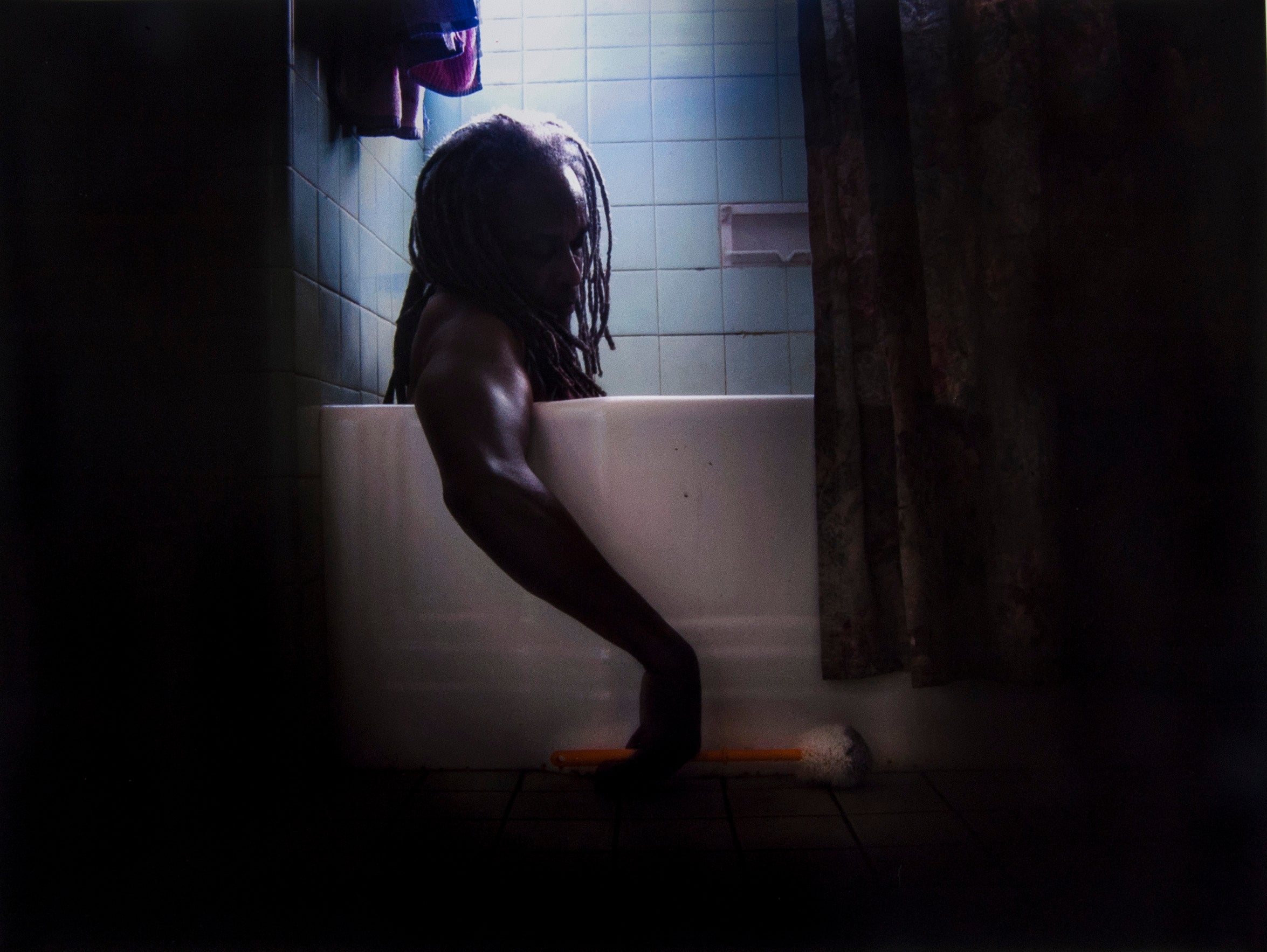 A man with dreadlocks sitting in a tub and reaching over for a brush on the floor. Very dark with blue tile.