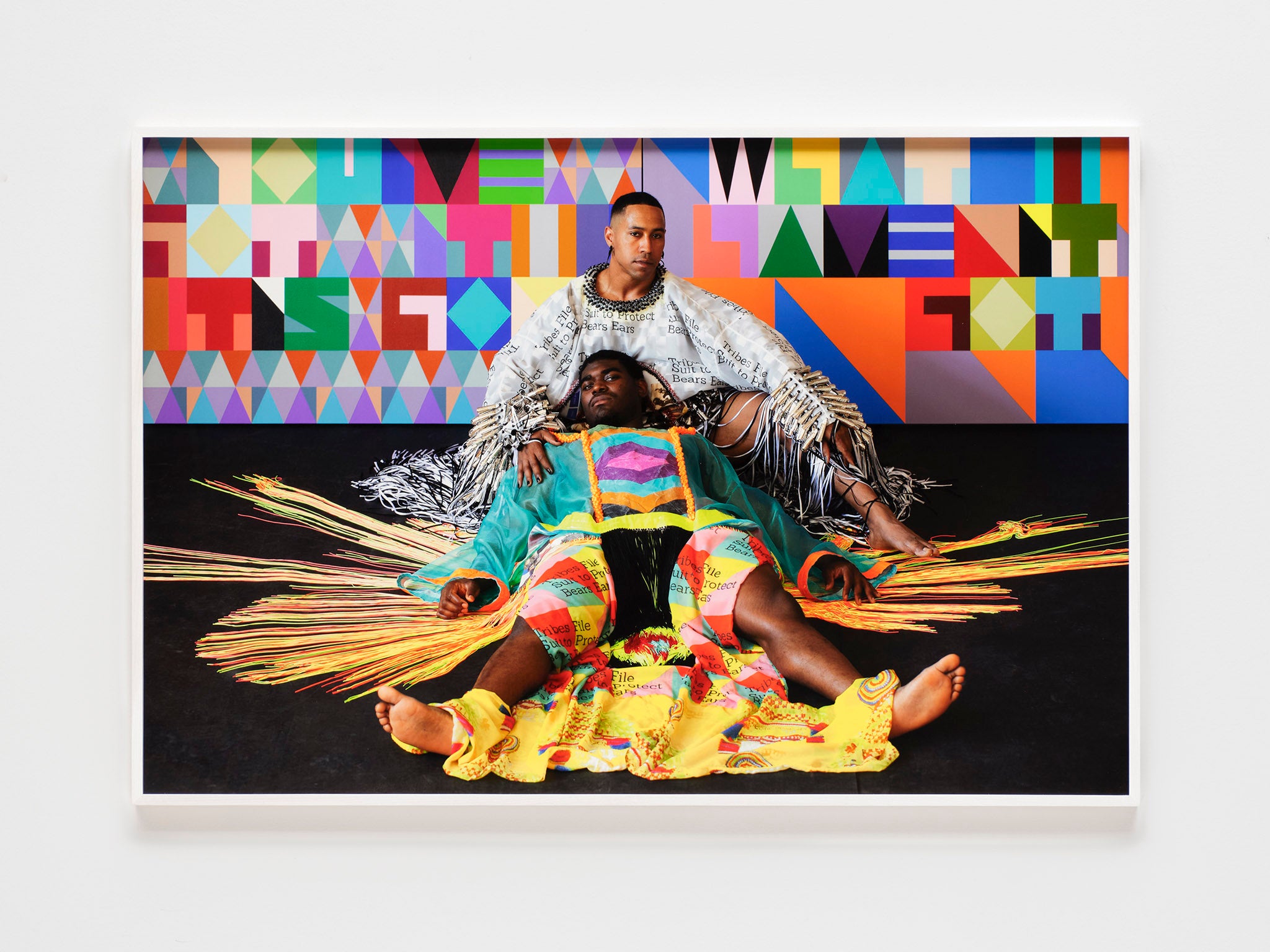 Photograph of two figures, dressed in rich brightly coloured fabric, sprawled on a dark floor in front of a wall of geometric patterns
