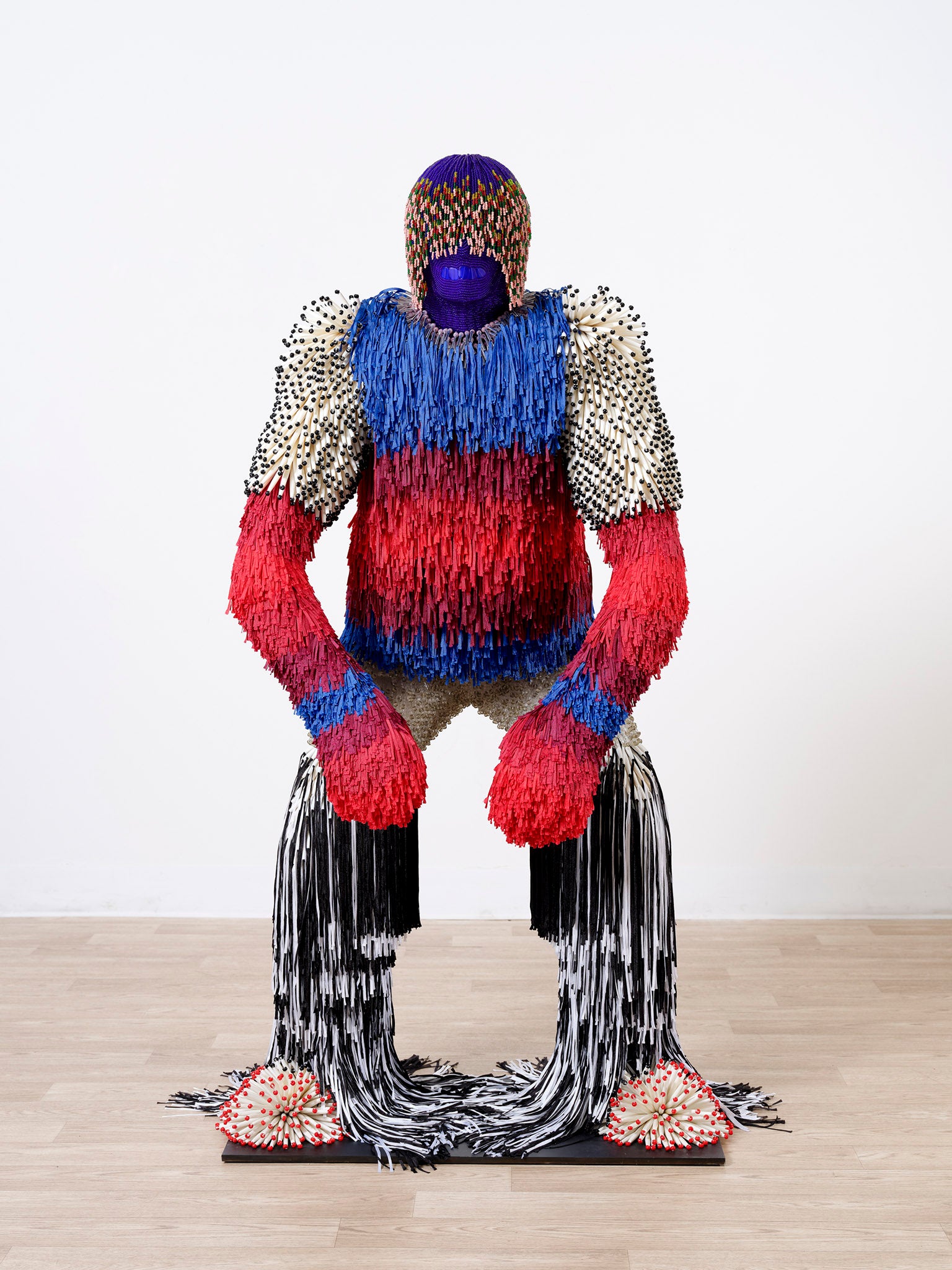 A humanoid figure in a semi crouch, made of brightly colored tassels and beads