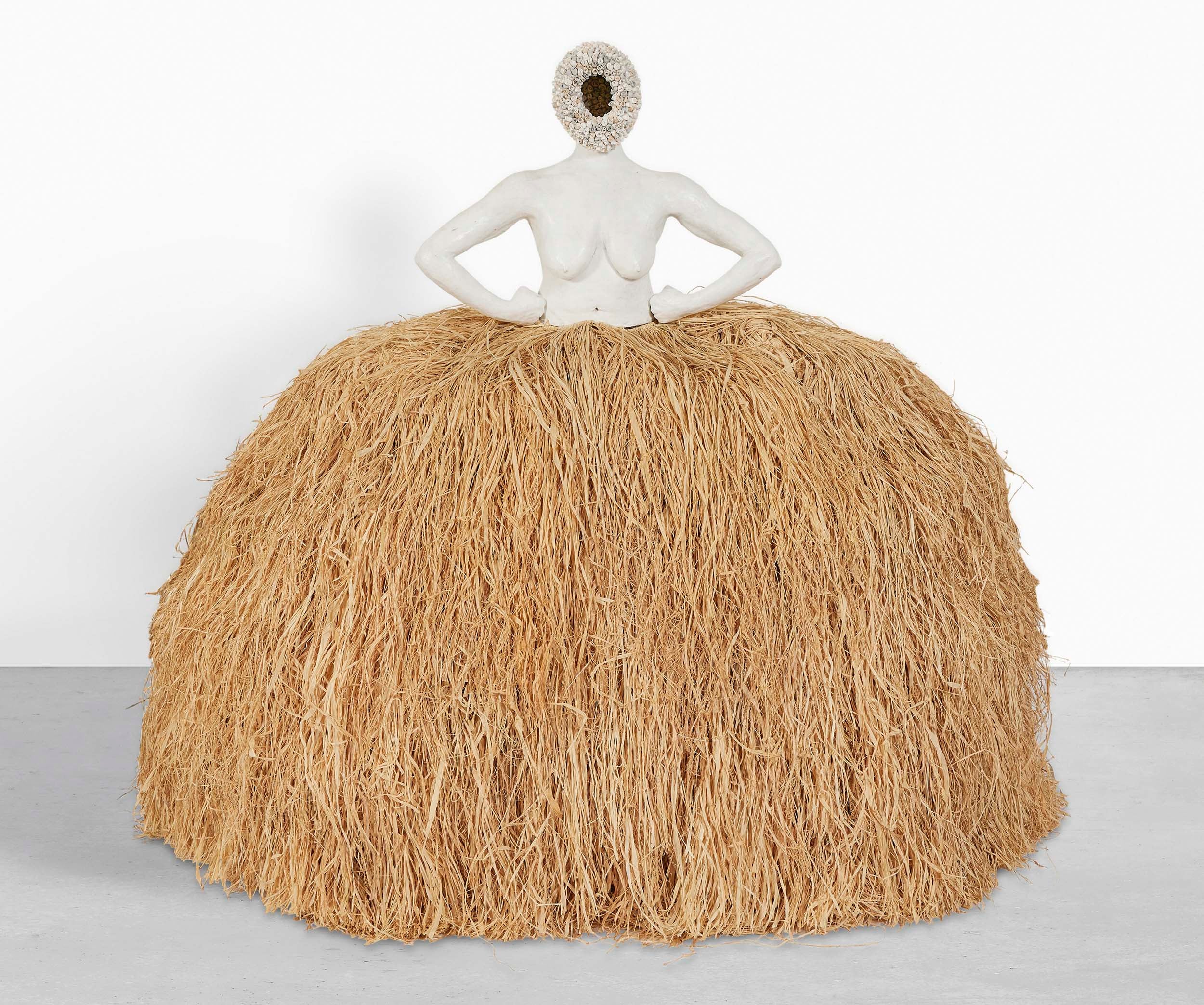 A statue of a faceless woman with her hands on her hips made of white material and whose head is a wreath of flowers. She is wearing a gigantic grass skirt much like the hoop skirts of the Victorian era.