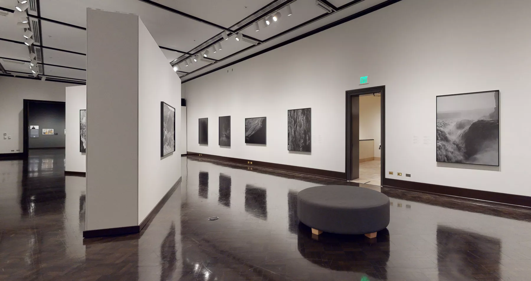 A white wall gallery with several black and white framed photos on the walls. There is a free standing wall to the left and a circular couch on the right.