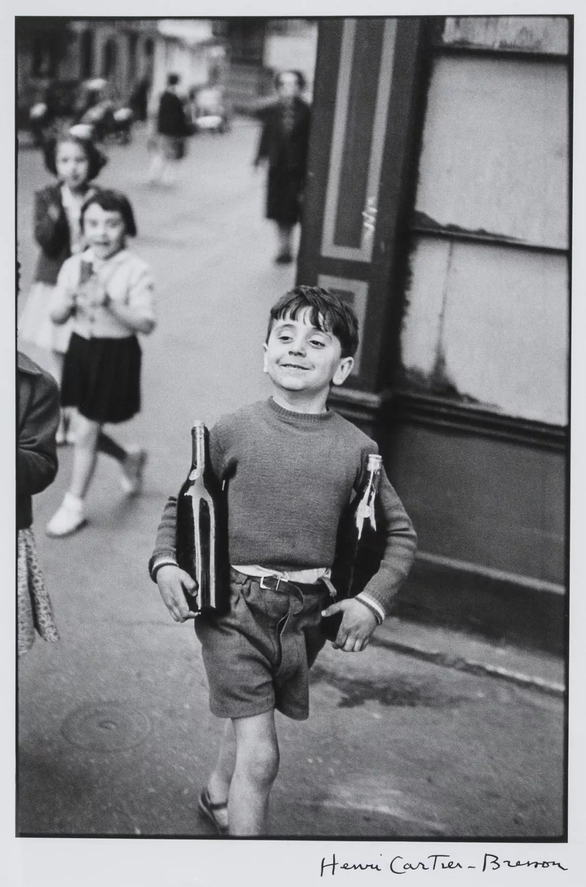 A black-and-white photograph of a young boy in a street, carrying two bottles. He has a proud expression and is surrounded by other children and adults in the background.