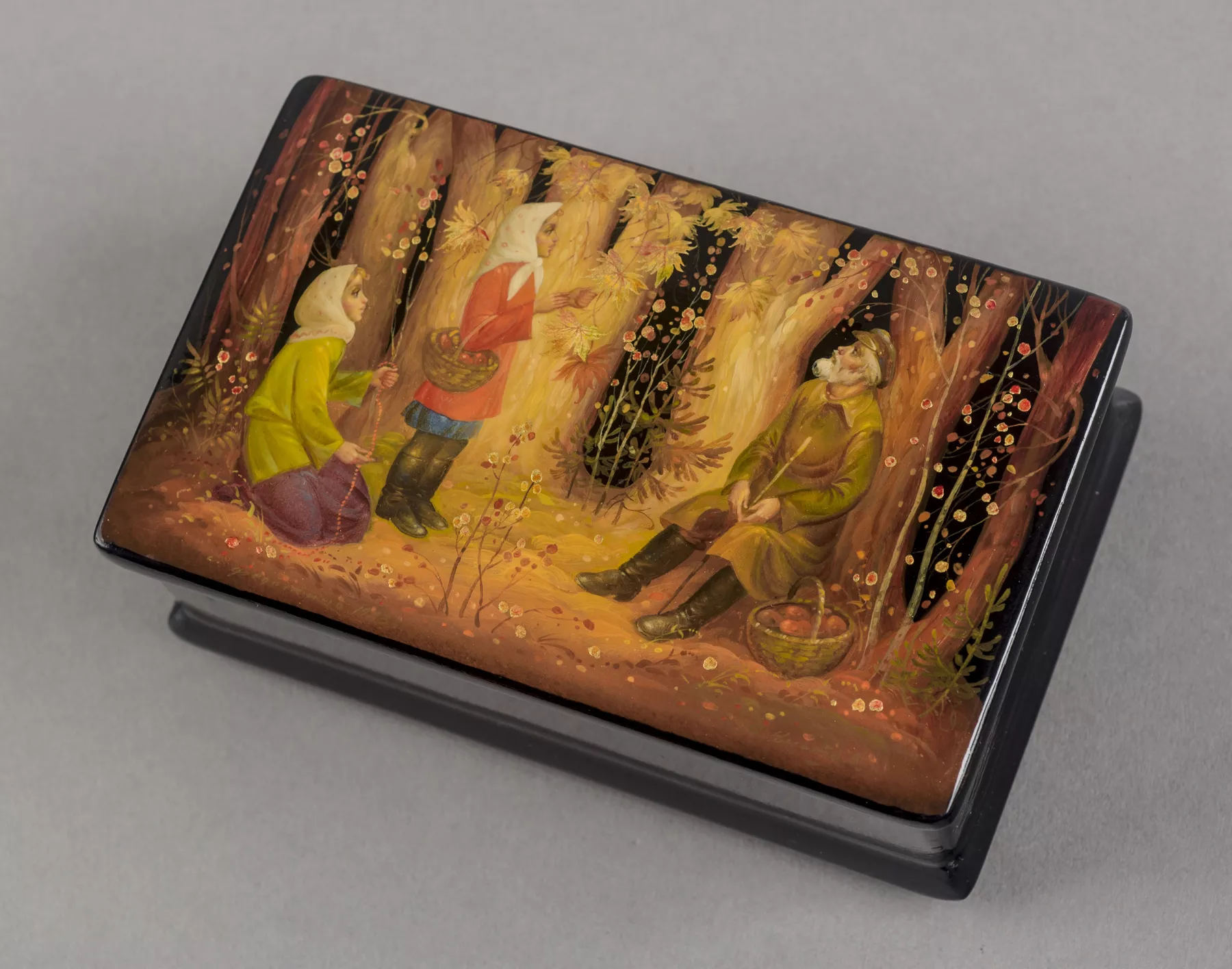 A painted Russian lacquer box showing a fairy-tale scene with three figures in a forest clearing, set against a dark background with trees and foliage, with rich autumnal colors.