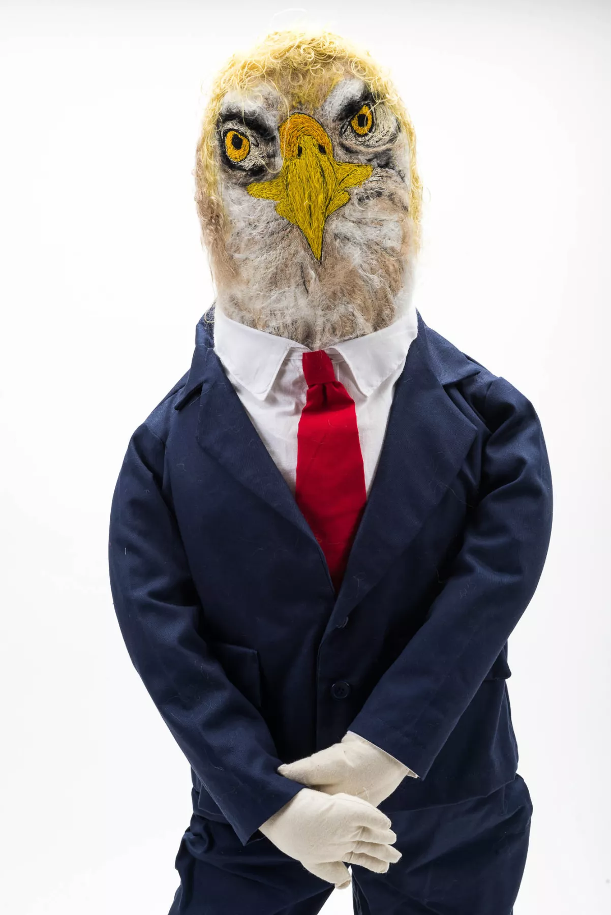 A sculpture of an eagle-headed figure dressed in a suit and red tie. The anthropomorphic figure has a serious expression, blending human and animal characteristics.