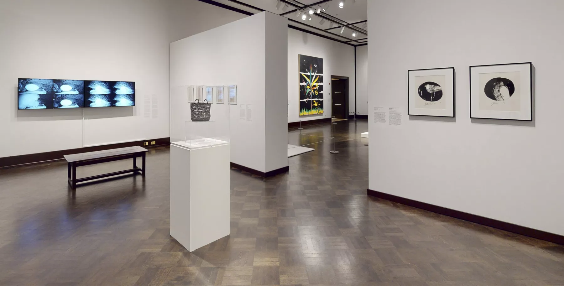 White walled gallery with several framed artworks and a video screen. There is a bench at the far end and a pedestal with an artwork in the center of the room. Through a doorway a glimpse of another gallery is visible.