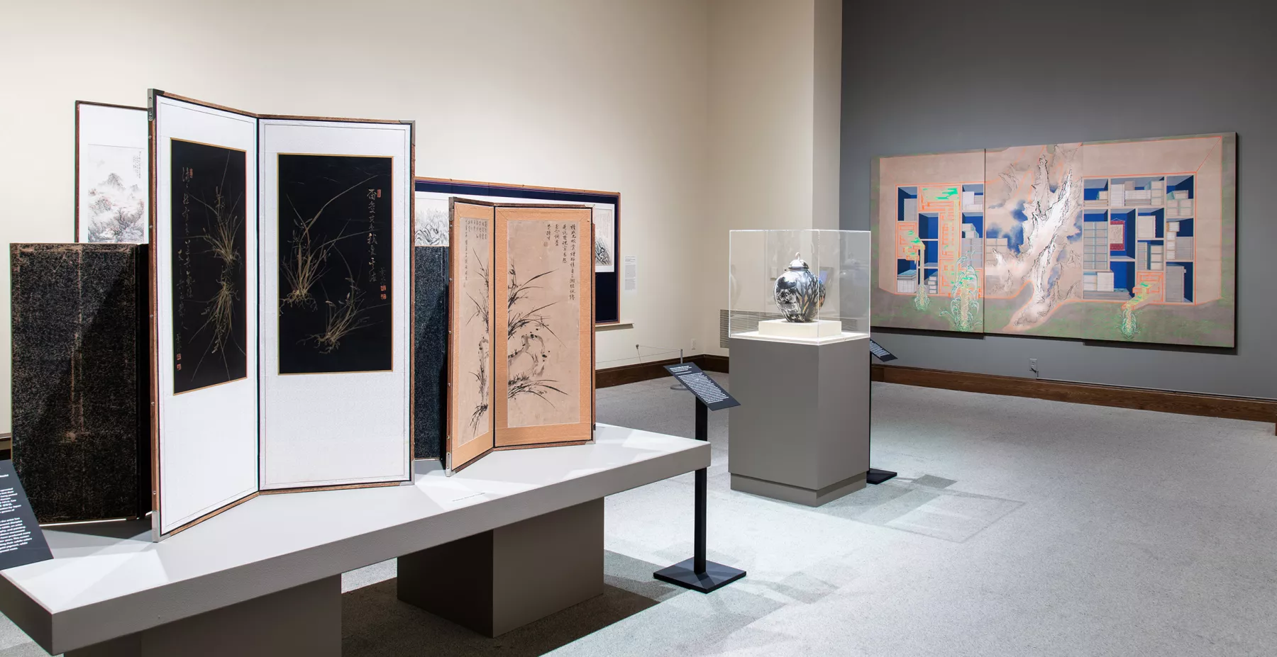 A gallery space with a painting on a grey wall, a ceramic pot on a pedestal and a table on the left with several Korean screens.