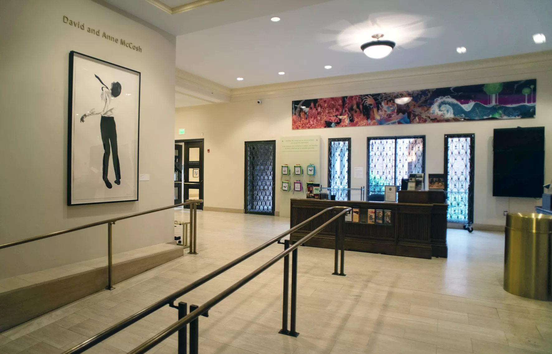Lobby of a museum with a welcome desk and a large framed print of a person dancing on the left wall. There is a colorful artwork over the desk and handrail through the middle of the image.