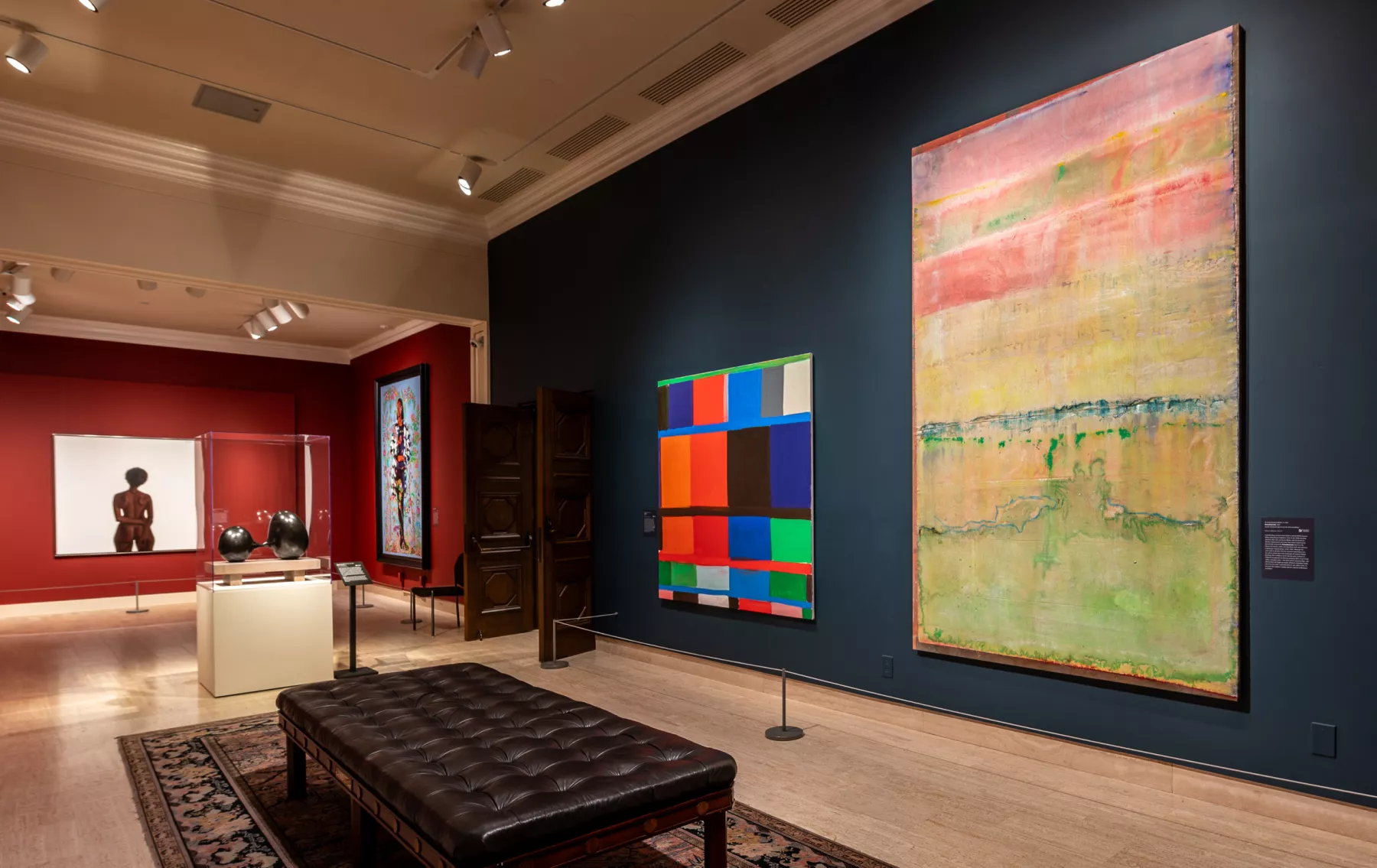 A gallery space with a red wall and a blue wall with several paintings. The two paintings on the right are very colorful abstract, while the two on the left are of abstract human figures and are more muted.