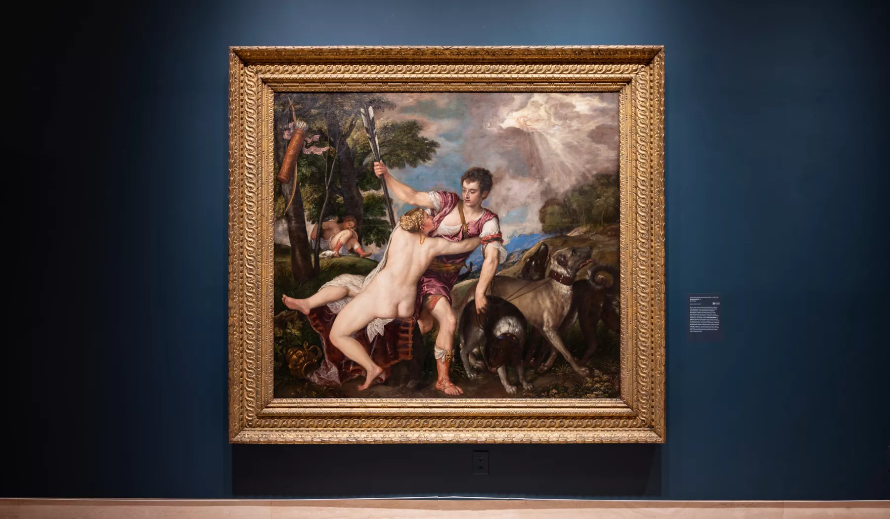 A large framed painting hangs on a dark blue wall. It has an ornate frame and the painting is of a hunter in medieval clothing with several hounds and a nude woman grasping him as if begging him not to leave.