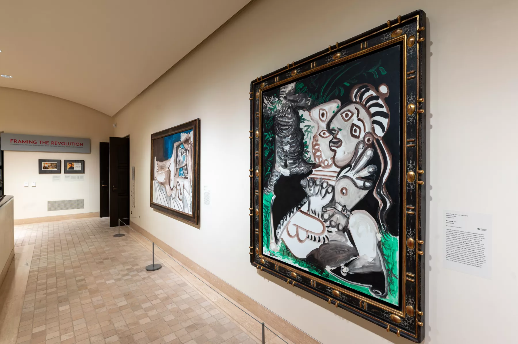 A hallway with two large Picasso paintings. The floor is tile and the view is from the side viewing down the hallway. The paintings are very large and the frames are ornate.