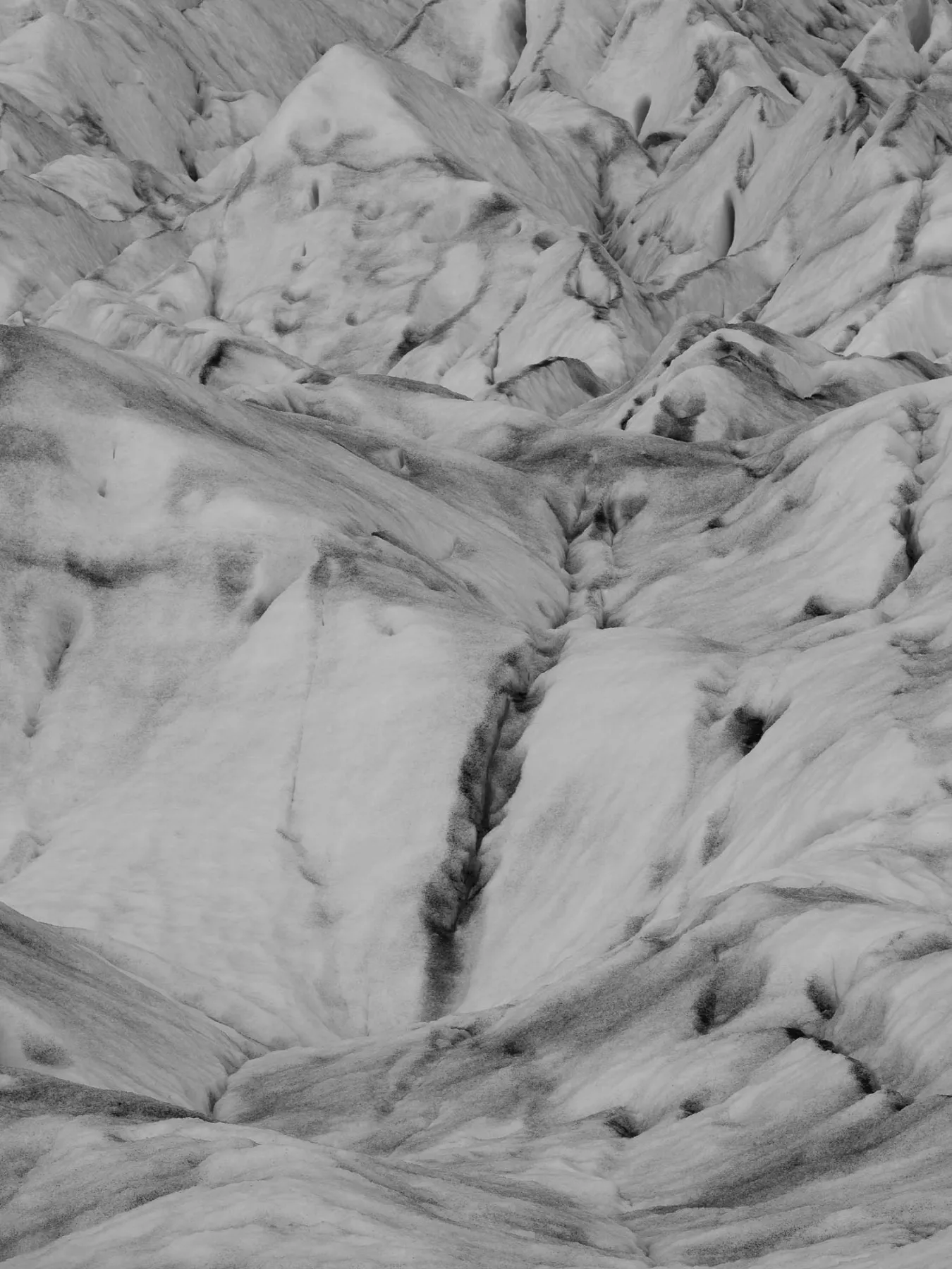 A monochrome photograph of a glacier's intricate, snow-covered surface with deep crevices and textured patterns.