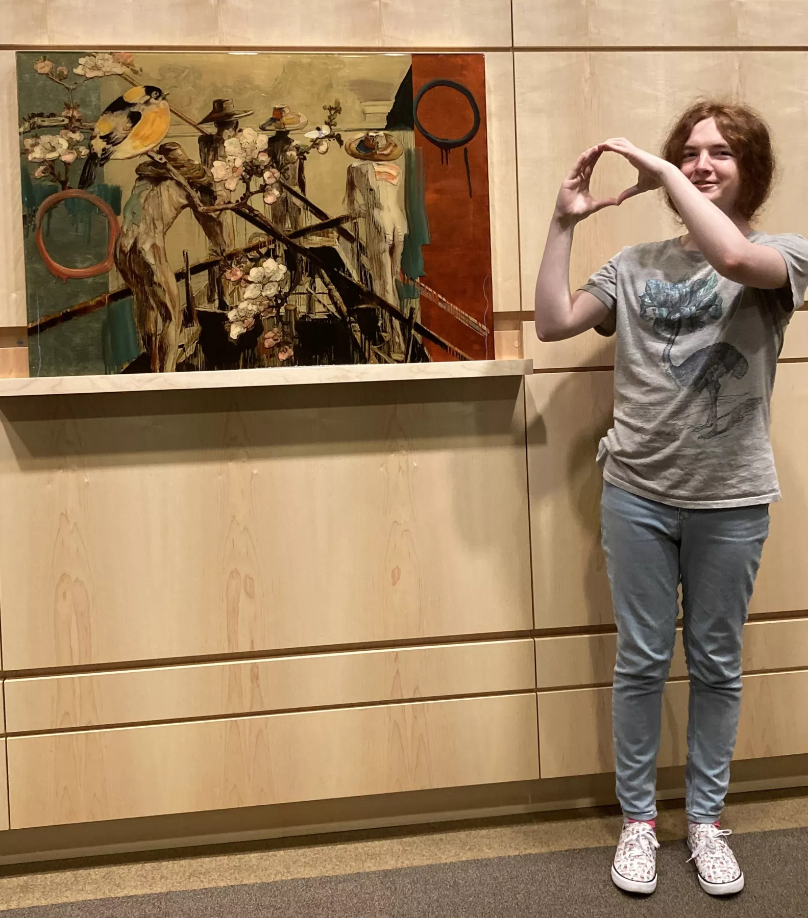 A person making a heart shape with their hands, standing next to a painting by Hung Liu titled 'Searching for Penglai,' which depicts a bird perched on a branch with a background of abstract circular and floral motifs.