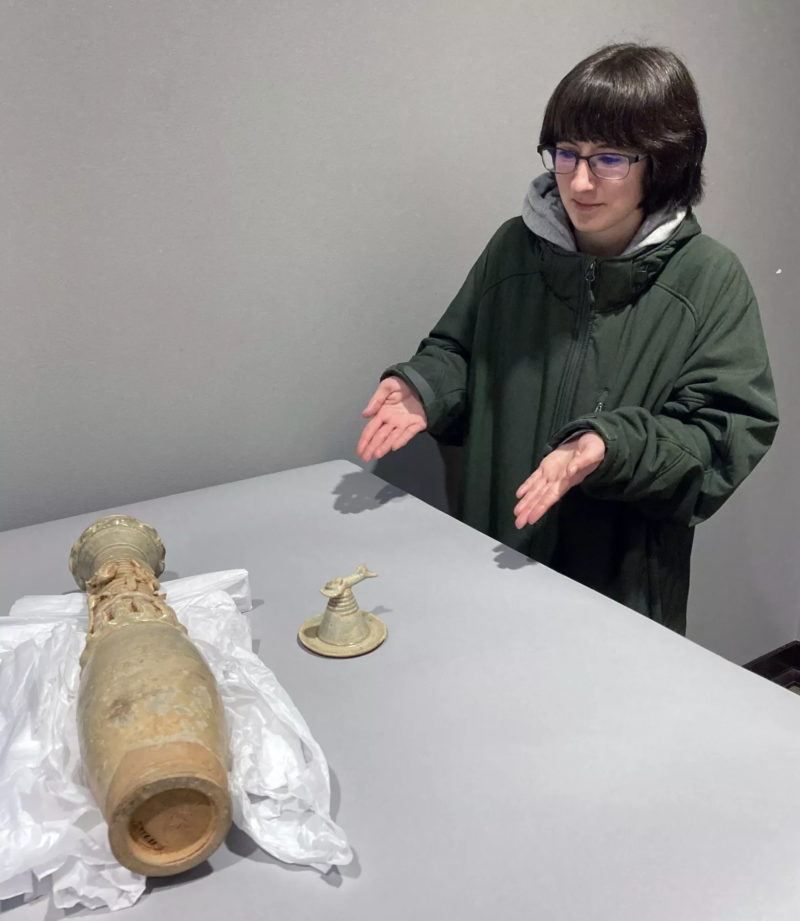 A person gesturing towards two ceramic artifacts, a large spirit jar lying on its side and a smaller, upright jar with a spout, both displayed on a table. The person is wearing a green coat and glasses.