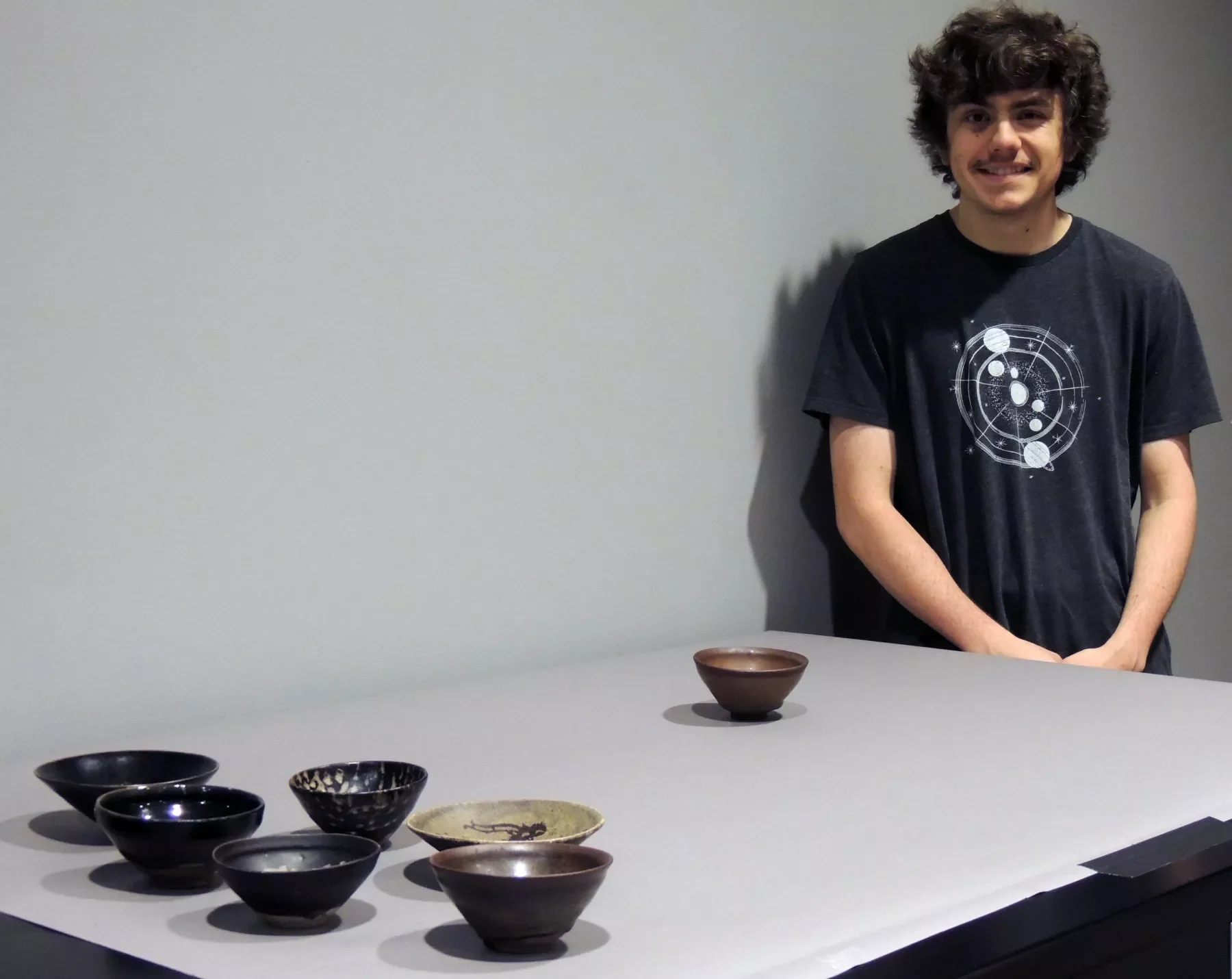 A person standing next to a display of seven tea bowls, arranged in a row, each bowl slightly different in shape and glaze color, ranging from dark to light tones.