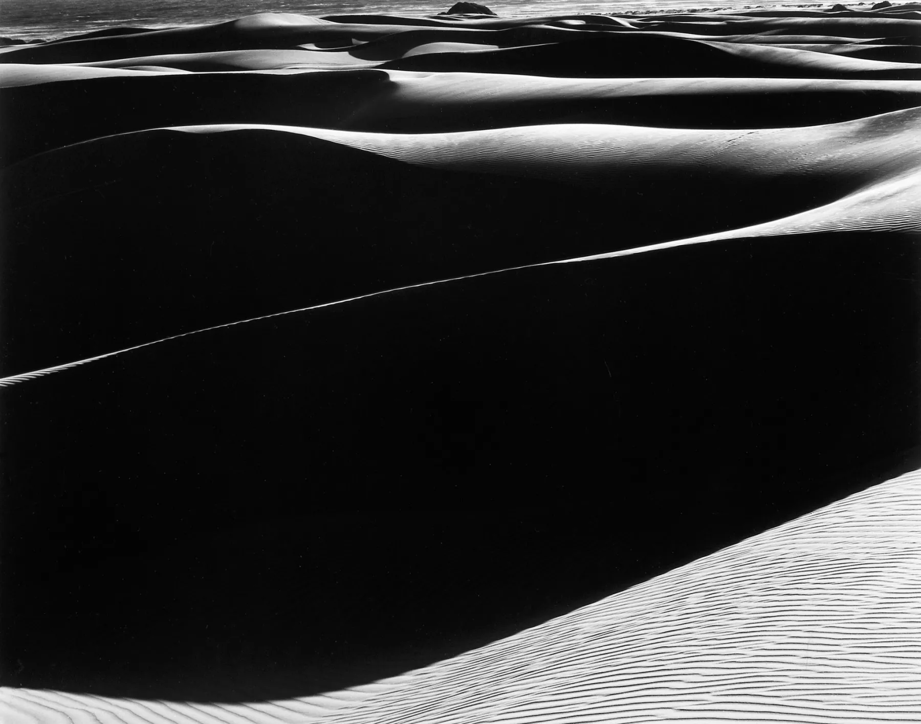A black-and-white photograph of sand dunes with smooth, flowing contours, casting deep shadows and highlights that create a striking contrast between light and dark areas. The textures of the sand and the play of light evoke a serene yet dramatic landscape.