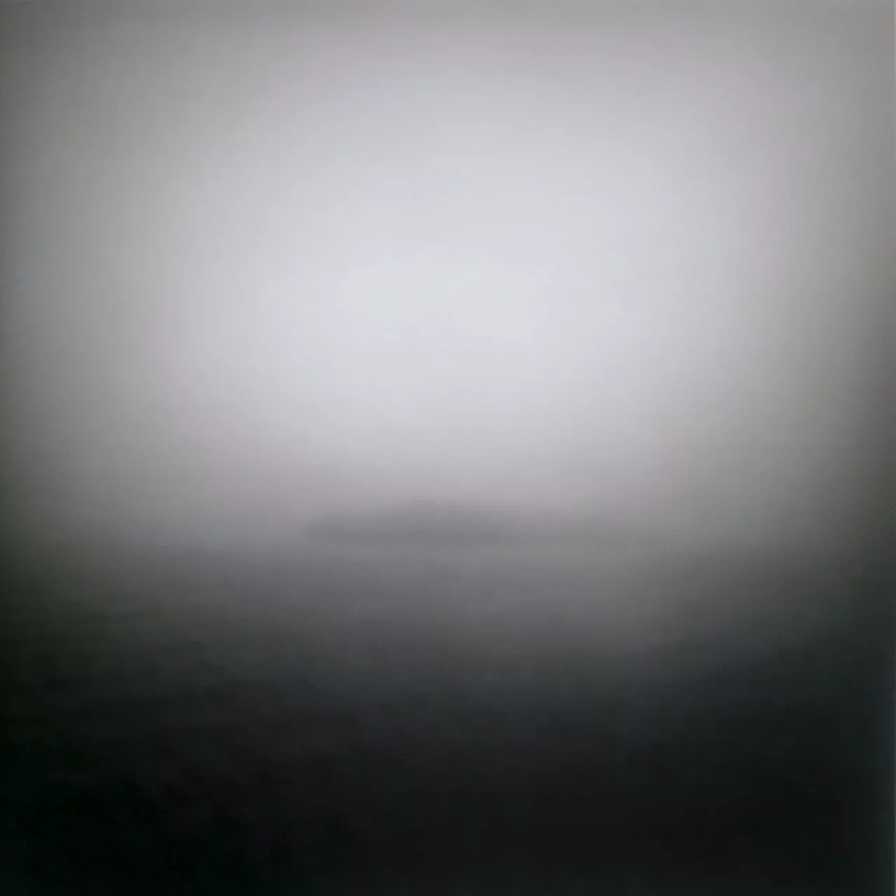 A minimalist, soft-focus photograph of a distant island shrouded in thick fog, almost blending with the horizon. The image has a dreamlike quality with muted tones, making the island appear as a faint silhouette against the misty surroundings.