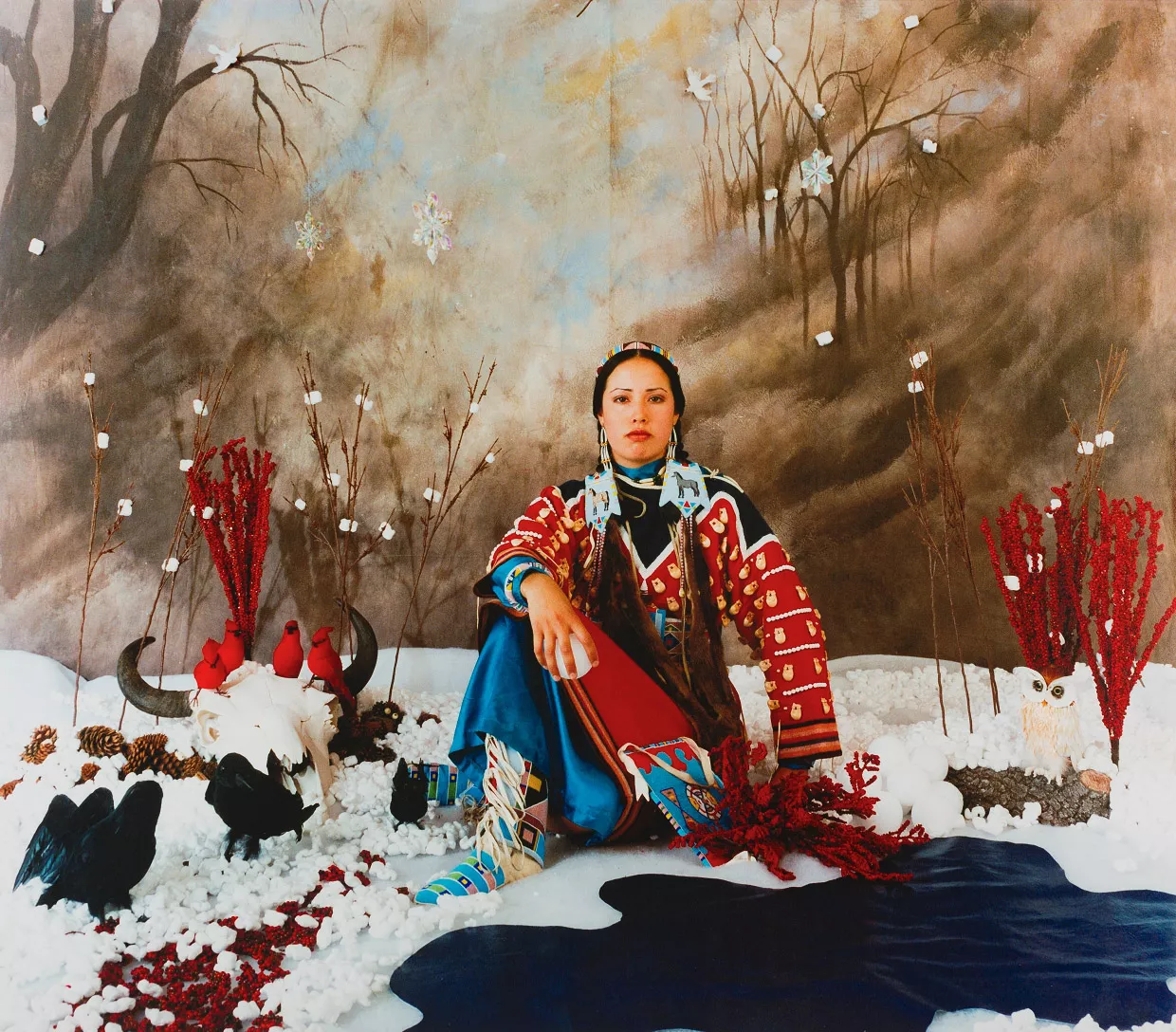 A vibrant color photograph of a woman dressed in traditional Indigenous attire, seated in an elaborately decorated indoor setting. She is surrounded by various objects including red branches, pine cones, and an owl figurine, with a painted backdrop depicting a wintery scene.