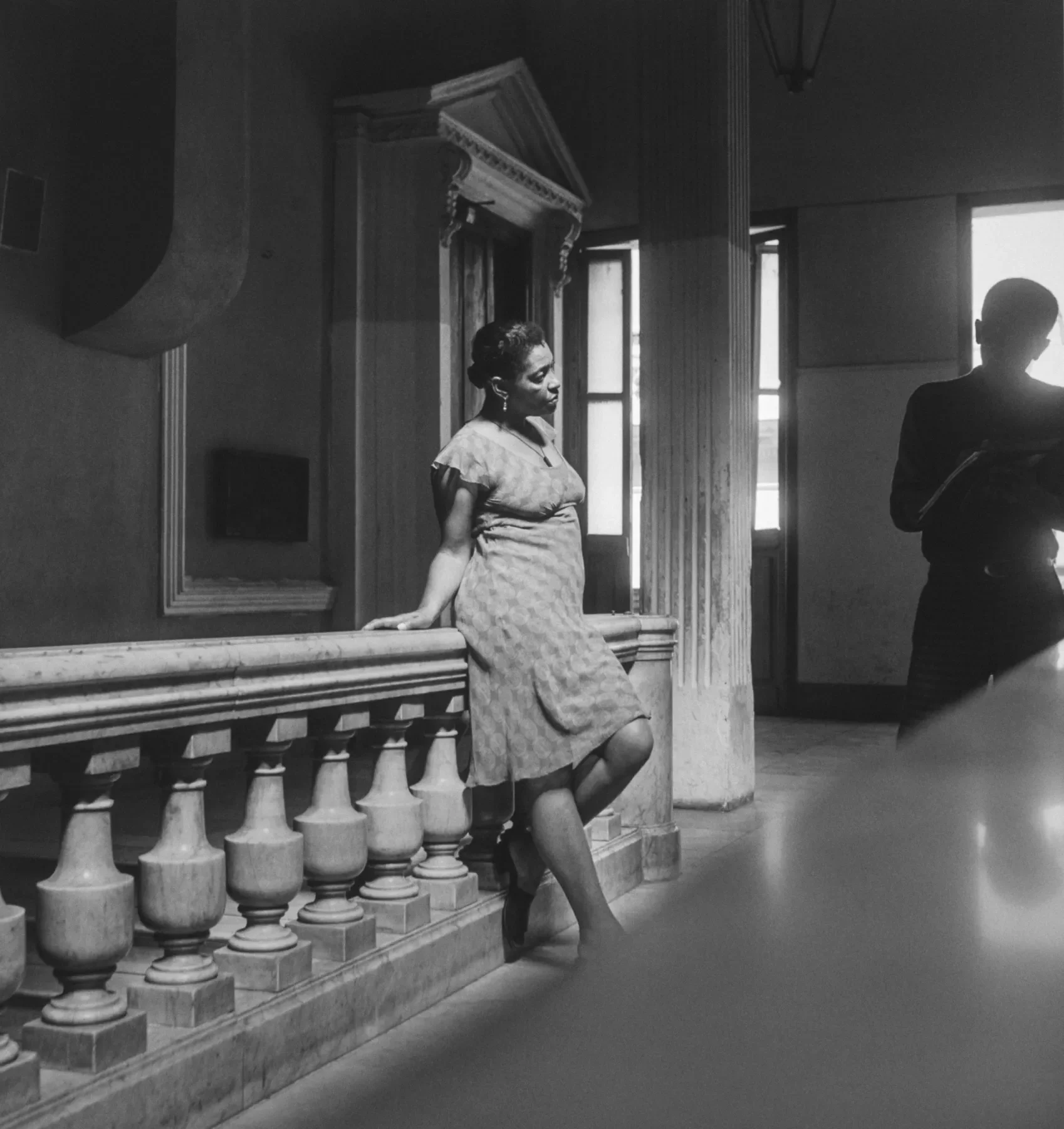 A black-and-white photograph of a woman leaning against a marble railing inside a grand, dimly lit building. She gazes off to the side, her posture relaxed, while another figure stands in the background, partially obscured by shadow.