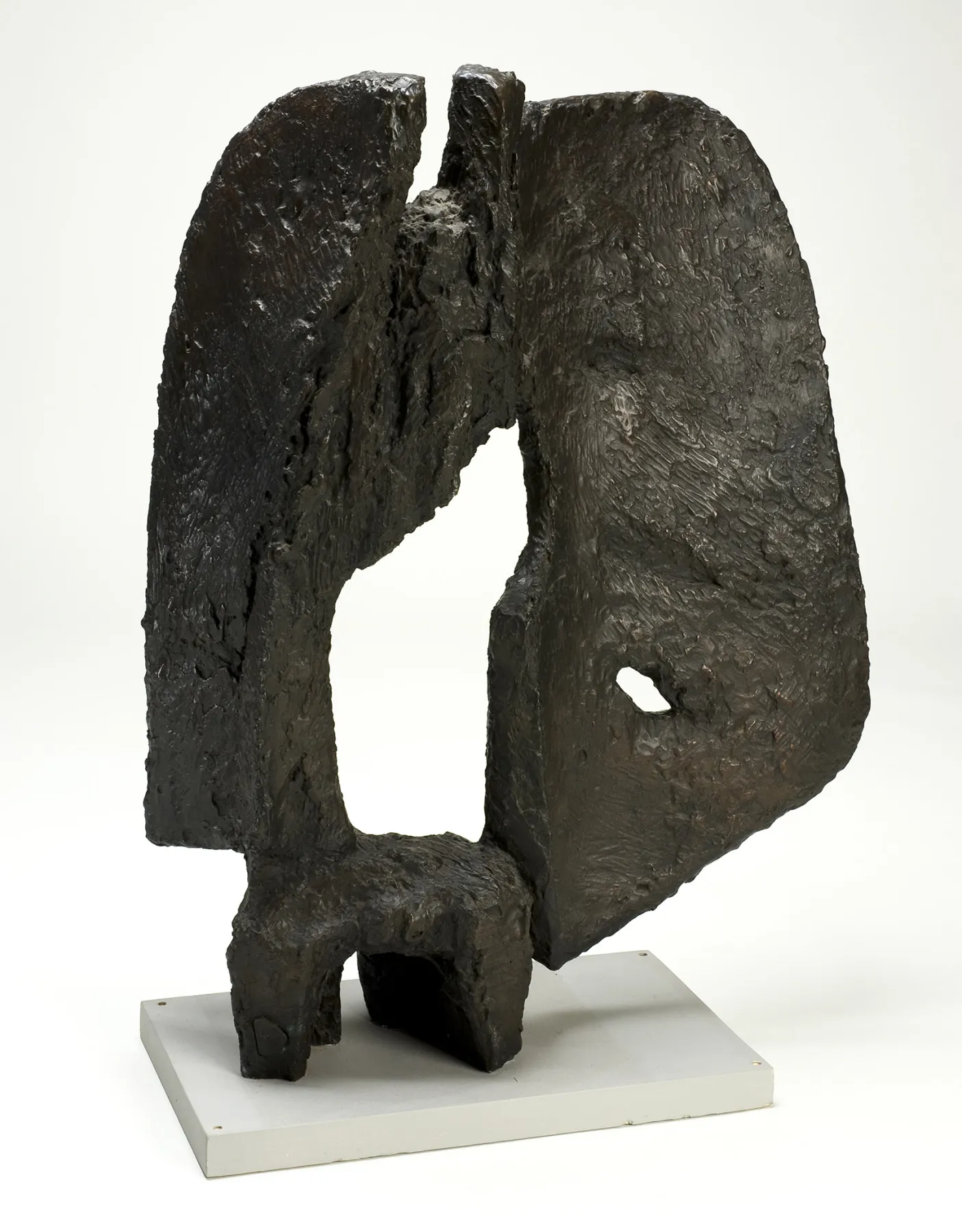 A dark bronze abstract sculpture with a rough, textured surface stands on a light-colored rectangular base. The piece has an irregular shape with two large, curved sections that frame a central void. One side features a small hole, adding an organic element to the composition. The sculpture appears weathered and has a dynamic, almost fragmented form, evoking a sense of movement or decay. The background is a neutral white, highlighting the dark patina of the metal.