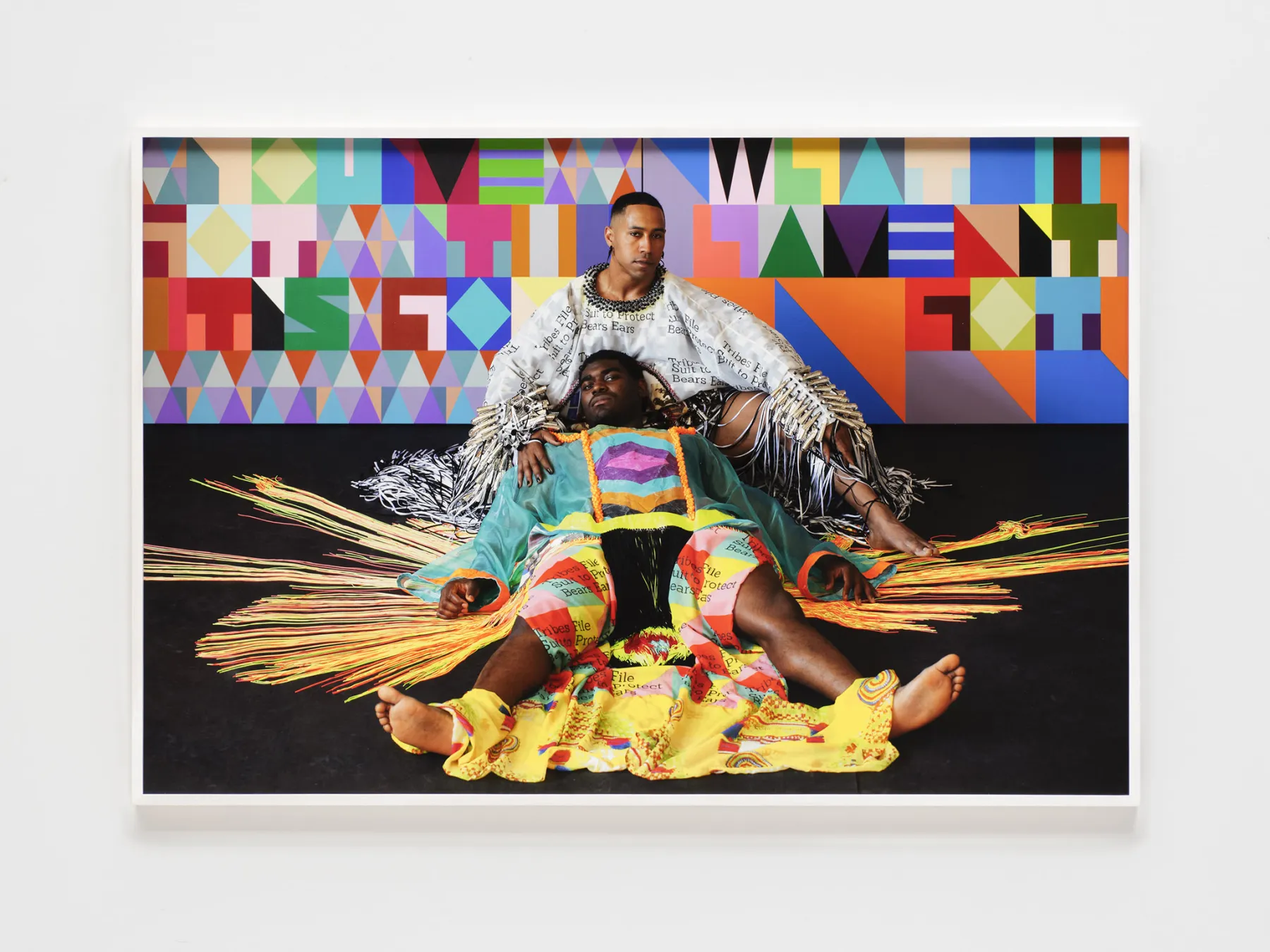 A photograph of two people posed within a vibrant, art-filled environment. One person sits against a wall covered in a bold, colorful geometric pattern, wearing a white garment printed with text and adorned with metallic silver fringe. A second person lies on the floor with their head near the first person’s lap, wearing a translucent green tunic over shorts also printed with text. Bright yellow and orange fringe-like materials radiate out from the central figures across a dark floor.