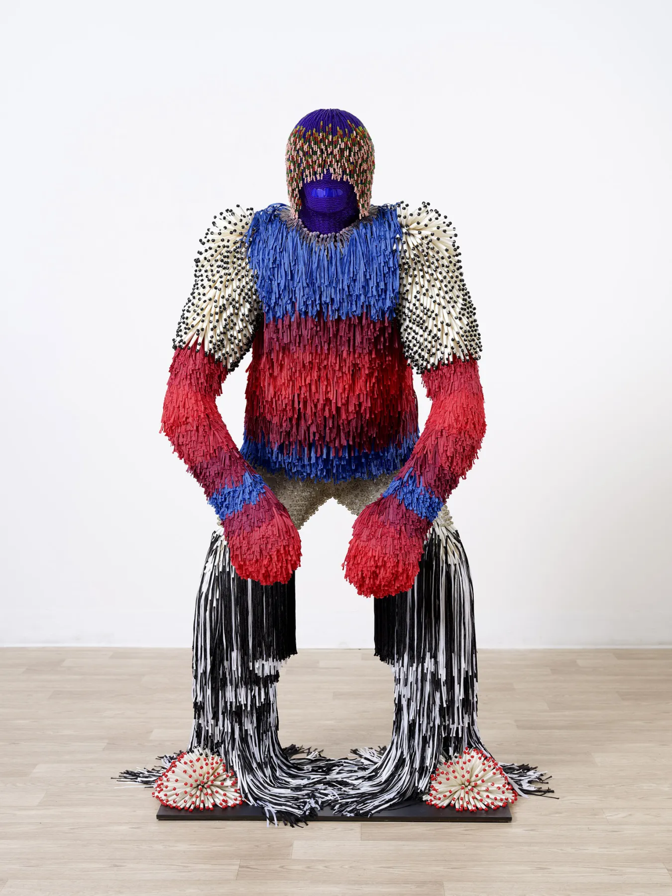 A large-scale, colorful sculpture of a humanoid figure standing in a gallery. The figure has a purple, textured head topped with a multicolored knit-style cap. Its torso is covered in layers of vibrant blue and red fringe. The shoulders are adorned with white, spiky protrusions tipped with black. Long black and white fringe flows down the legs, pooling onto a black square base on a light wood floor. The "feet" are represented by starburst patterns of red and white tipped spikes.