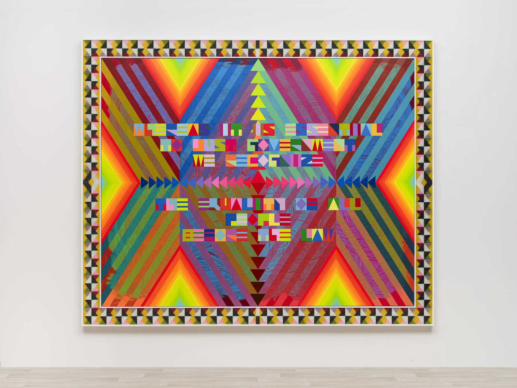 A rectangular artwork mounted on a white wall featuring a vibrant, geometric "radiating" pattern of concentric diamonds in rainbow colors. Centered on the piece is stylized block text in various colors that reads: "WHEREAS IT IS ESSENTIAL TO JUST GOVERNMENT WE RECOGNIZE THE EQUALITY OF ALL PEOPLE BEFORE THE LAW." The piece is framed by a border of black, white, and yellow checkered triangles.