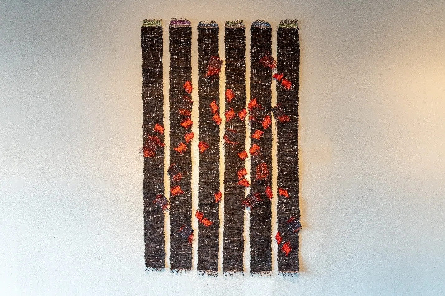 A textile art piece hanging on a white wall, composed of six vertical woven panels of dark, textured brown fabric. Scattered unevenly across the panels are numerous small, irregular patches of vibrant orange and red fiber pieces, giving the impression of floating autumn leaves or glowing embers drifting across a dark background.