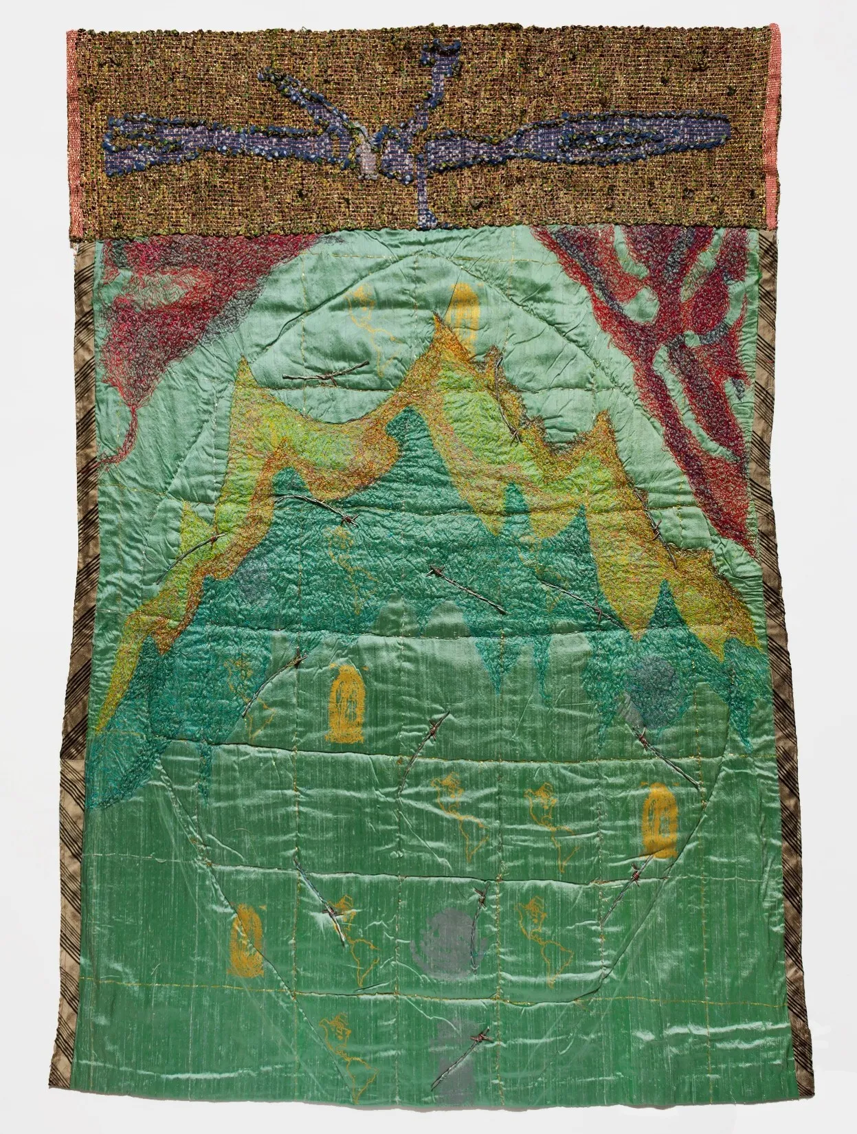 A vertical mixed-media textile art piece. The top portion features a wide, rectangular band of coarse, woven golden-brown fiber decorated with a large, abstract blue and purple shape reminiscent of a bird or an insect. The main lower section consists of a quilted, shimmering green fabric stitched with grid lines. Layered over the green backing are embroidered yellow outlines of continents, abstract shapes resembling yellow footprints, and a large, textured mountain-like form in shades of green and yellow that dominates the upper half, framed by dark red branch-like patterns in the top corners.
