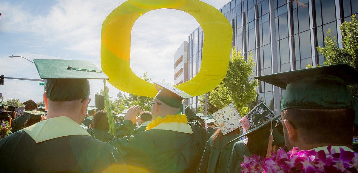 A group of people in green caps and gowns hold a large yellow "O" in the air