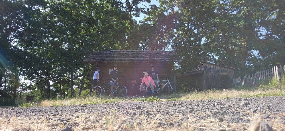 Group of people out by a shed, with a gravel road, in 4 people with bikes
